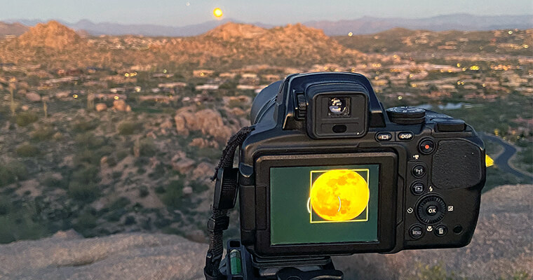Moon Hike Camera on a tripod capturing a full moon over a rugged landscape at twilight.
